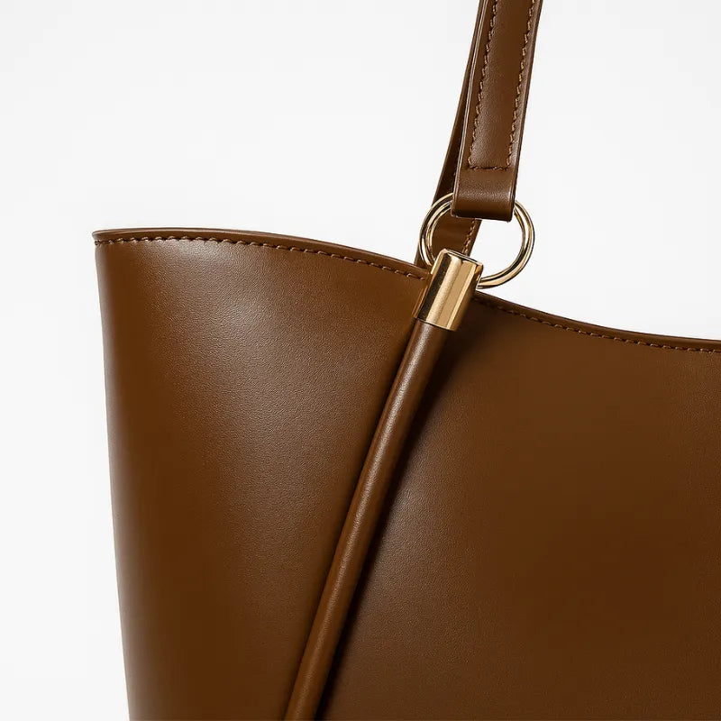 Detalhe da lateral da bolsa de ombro marrom. A imagem em close aproxima os detalhes metálicos e o material em Couro PU.