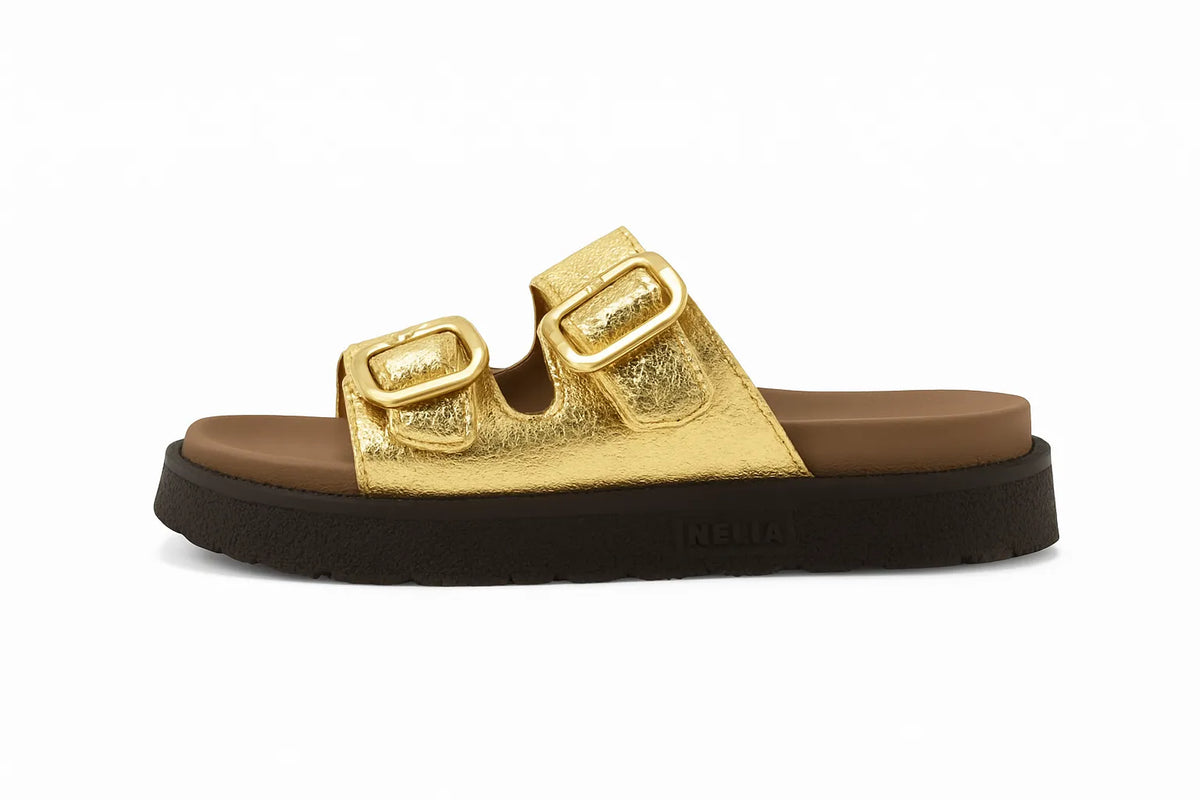 Sandália feminina dourada com fivelas, vista lateral em estúdio. A foto destaca o design flatform, o solado preto e as duas fivelas ajustáveis na parte superior.