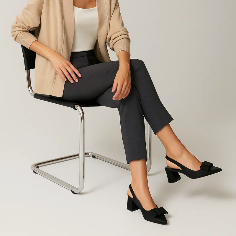 Modelo usando o scarpin slingback preto com laço da Rozze, sentada. A imagem mostra o calçado em uso, complementando um look de trabalho com calça social e blazer.
