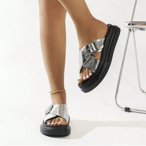 Modelo usando a papete feminina prata de fivela da Rozze, sentada. A imagem mostra o calçado em uso, complementando um look casual e realçando o conforto e a versatilidade.
