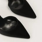 Scarpin preto salto alto fino em close no bico, tiras ou fivela, com textura e metal em destaque, sobre fundo claro