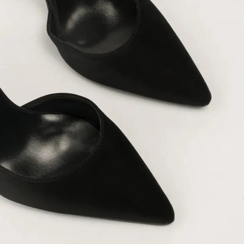 Scarpin preto salto alto fino em close no bico, tiras ou fivela, com textura e metal em destaque, sobre fundo claro