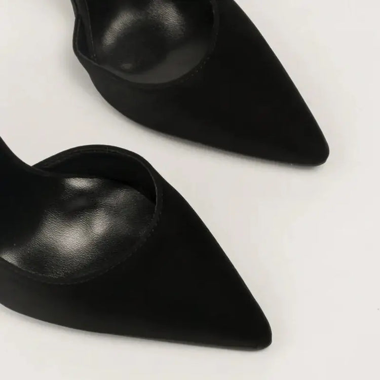 Scarpin preto salto alto fino em close no bico, tiras ou fivela, com textura e metal em destaque, sobre fundo claro